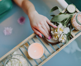 A hand with pink nails touches flowers on a bamboo tray with spa items.